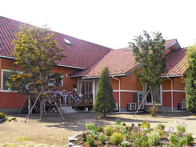 朱色の屋根の建物と庭 外観 庭に面した木陰でゆったりとした時を過ごせる環境です。日差しを避けながら穏やかに交流する方々の姿が見られます。