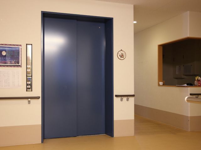 青い扉のエレベーター 廊下 手すりが設置された清潔感のあるエレベーター前です。安全に配慮された設計で、快適な移動をサポートします。