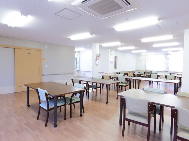 広々とした明るい食堂 食堂兼機能訓練室 ゆったりと配置されたテーブルと椅子が整然と並べられ、日中の自然光が差し込む心地よい食堂です。機能的な空間での仕事をご検討なら、この明るい環境は働く方にとっても居心地が良いでしょう。