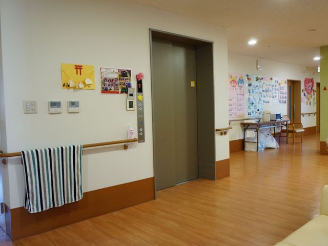 多機能な廊下 エレベーター 手すりの付いた廊下は、ゆったりとした歩行スペースを確保し、壁面には明るい色の工作物が飾られています。安全かつ快適な移動を支援する設計です。