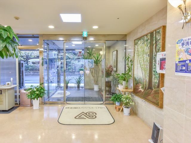 明るいエントランス風景 入口  清潔で開放的なエントランスは、植物に囲まれ安らぎの空間を提供しています。来訪者を暖かく迎え入れる場所となっています。