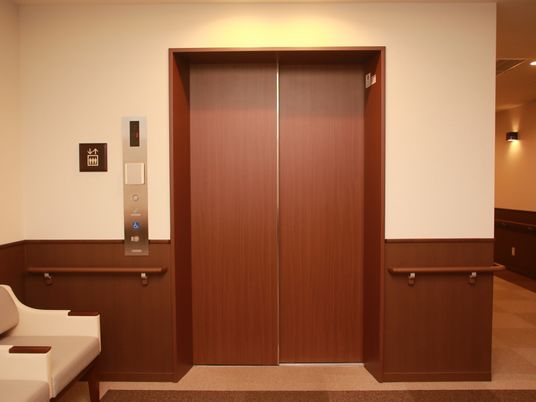 木目調のエレベーターの扉の前にソファが設置されている。エレベーターの扉、通路の腰壁、手すりが濃い茶色で、落ち着いた雰囲気の空間になっている。