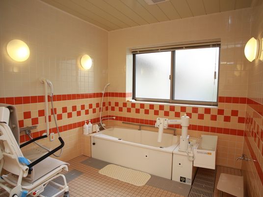 暖色系のタイルが張られた明るい雰囲気の浴室に、大きな四角い浴槽と、車椅子型の入浴用チェアが設置されている。