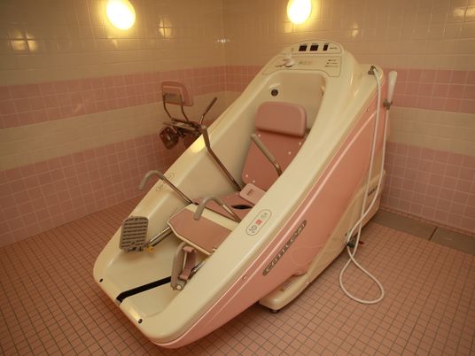 広いタイル張りの浴室に、車椅子タイプの入浴装置が設置されている。浴室の隅に、予備の椅子が置かれている。