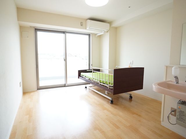 明るい居室のベッドと洗面台 居室 広々とした居室には、日当たりの良い窓と必要な設備が整っており、プライバシーもしっかり保たれています。