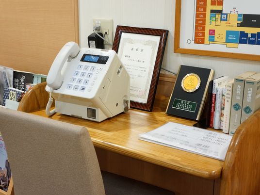 木製のテーブルの上には大きな電話が置かれている。賞状や本などもある。壁には施設構内図が貼られている。