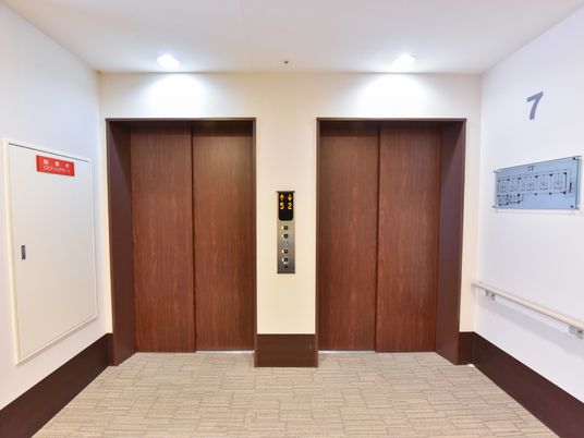 白い壁に茶色のエレベーターの扉がマッチしている広々としたエレベーターホール。2基設置されているので、ゆとりをもって利用できる。