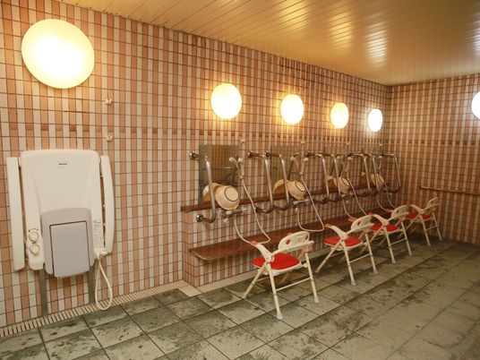 広いタイル張りの浴室に、シャワーが４つ並んでいる。鏡の両側にステンレス製のグリップが施され、洗い桶や入浴用の椅子が置かれている。