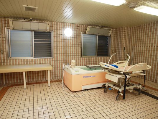 窓があるタイル張りの広い浴室に、ステンレス製の器具がついた長方形の浴槽と、可動式のストレッチャーが置かれている。