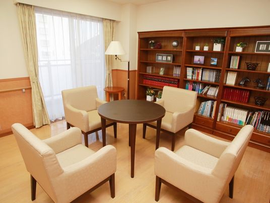 円形のテーブルを囲むように一人掛けソファが４脚置かれている。背の高い木製の棚に、書籍類や小物が並べられている。