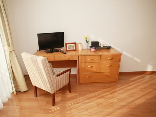 高さの揃った木製の机と収納家具が並べて設置されており、その上にテレビや小物が乗せられている。エレガントなデザインの椅子が置かれている。