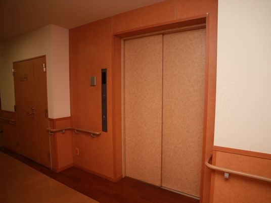 暖色で統一された空間で、木の温もりを感じられる内装である。エレベーターの扉があり、壁には手すりが連続している。
