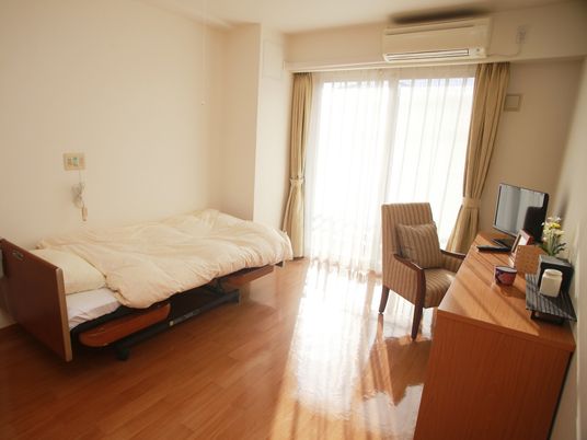 掃き出しの窓がある明るい部屋に、ベッドが据えられている。手前には、木製デスクと布張りの肘掛け椅子が置かれている。