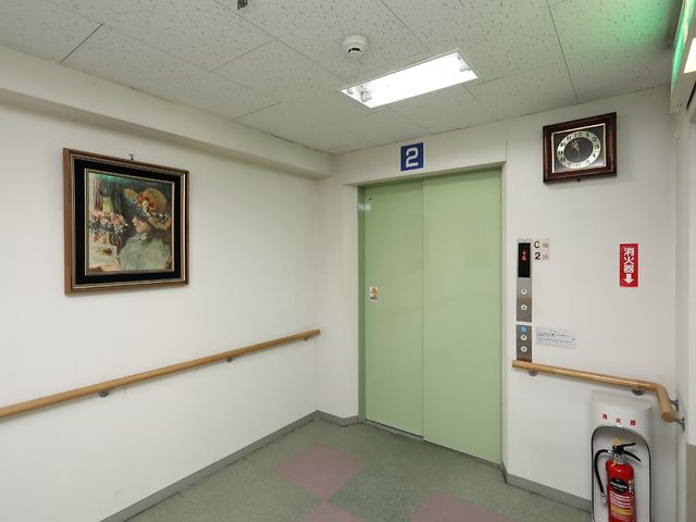 廊下と緑の扉の壁面 エレベーター 清潔感のある廊下は、壁面にアートが飾られ落ち着いた雰囲気を演出しています。手すりも設置され安全性にも配慮されており、快適な職場環境が整っています。