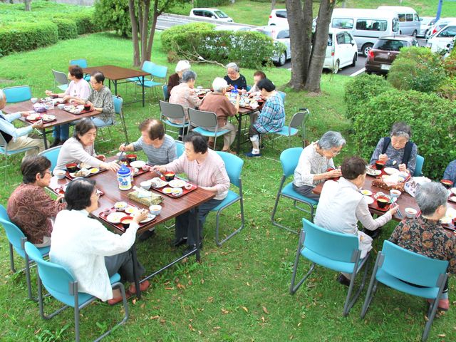 屋外で楽しむ食事会 緑あふれる中庭で、利用者同士が和やかに食事を楽しんでいます。心地よい風と日差しの中、コミュニケーションが生まれる空間です。