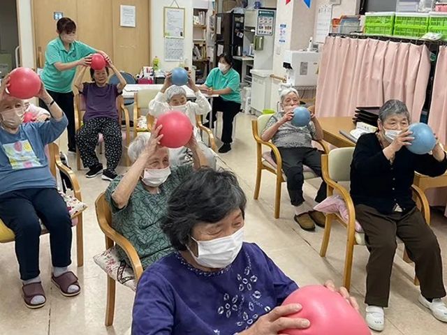 リハビリ中の利用者たち 機能訓練室 利用者の皆さんがリハビリに励んでいます。椅子に座って行う運動で、安全性が確保されています。
