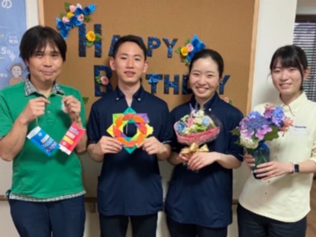 スタッフ集合の室内 内観   スタッフが並んで立ち、笑顔で花束や飾りを手にしています。明るく親しみやすい雰囲気で、働く人の様子が伝わります。