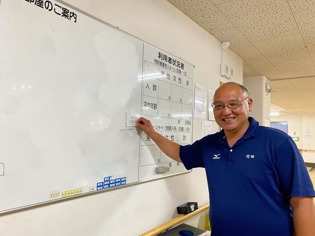 スタッフが掲示物を指差す様子 掲示物 ケアのスタッフが明るい表情で情報掲示板を指しています。こちらの施設では、透明感のあるコミュニケーションを大切にしています。