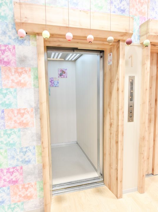 施設の写真 多彩な色を使った壁に小さな提灯が印象的な木目ドアのエレベーターで居室フロアなどに移動することができる。