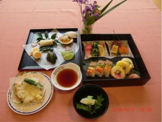 重箱が開けられ、本体と蓋にそれぞれ華やかな料理が盛られている。手前にはお吸い物と、丸い皿に載せられた天ぷらがある。
