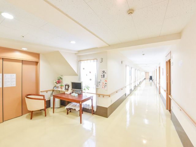 清潔感のある明るい廊下 施設内廊下 広々とした廊下は清潔で、掲示物や椅子が配置され、ご高齢の方々の交流の場となっています。デスクにはコンピューターもあり、情報の管理も行われている様子です。