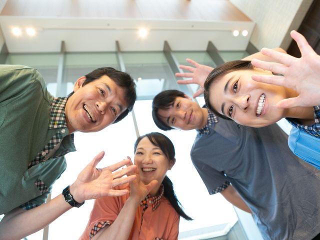 笑顔のスタッフ集合 スタッフイメージ   明るい表情のスタッフが集まり、手を振って親しみやすい雰囲気です。働く人同士の連携のよさが伝わります。