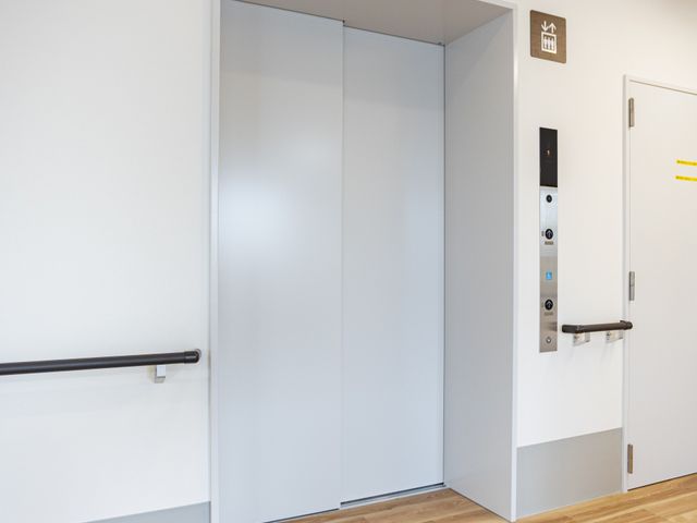 明るいエレベーター前の廊下 廊下   木目調の床と白い壁で、すっきりした印象の廊下です。手すりがあり、移動しやすい環境に見えます。