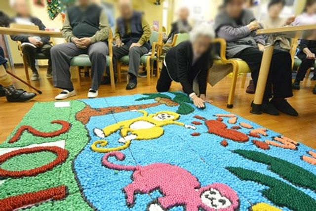 明るい作品展示の部屋 多目的室 手作り作品が床に広げられ、入居者たちが楽しそうに鑑賞しています。活気のある交流の場です。
