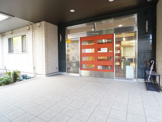 明るく広々としたエントランス 玄関 温かみのある赤い入口扉が迎える、清潔感溢れるエントランスです。通りから直接繋がる明るい通路が気持ちを和ませます。
