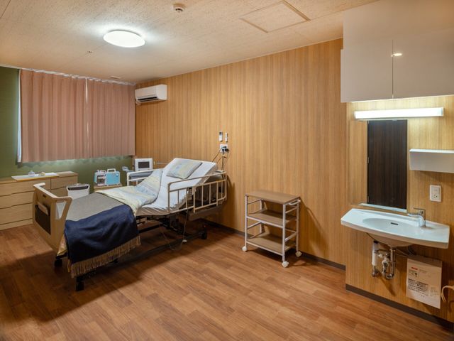 洗面台付きの居室 居室   木目調で落ち着いた雰囲気の居室です。ベッドと洗面台があり、ゆったり過ごしやすい環境に見えます。