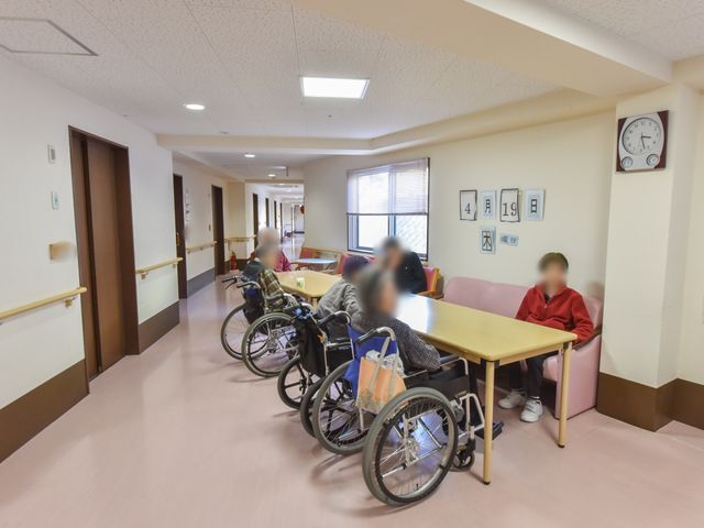 廊下にいる人々 談話コーナー  ゆったりとした廊下には椅子やテーブルが配され、休憩中の方々がくつろぐ姿が見受けられます。共に過ごす時間を大切にする環境が整っております。