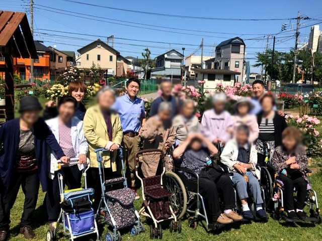 笑顔の集団が集う庭 庭 青空の下、多くの人々が庭で車椅子に座る方々と楽しそうに交流しています。自然に囲まれたリラックスできる場所です。