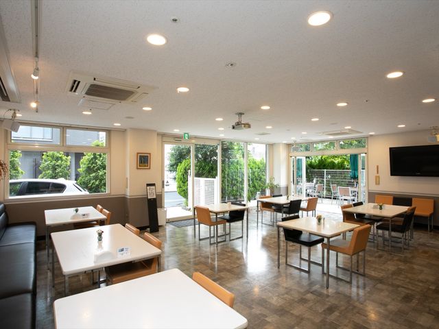 広々とした明るい食堂 カフェスペース ゆったりとしたダイニングルームは、清潔感があり、窓から自然光が差し込んでいます。テレビや外の緑も楽しめ、居心地の良い食事の時間を過ごせそうです。