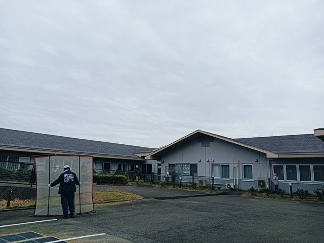 建物前の屋外 外観   平屋の建物と広い駐車スペースが見え、出入りしやすい印象です。周囲で作業する方の様子もあり、日中の落ち着いた環境です。
