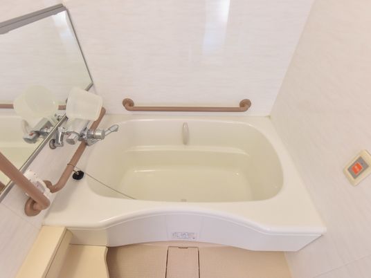 白色の浴槽の中や外周りに、手すりが設置されている。壁には横に長い鏡と緊急時呼び出しブザーが付いている。