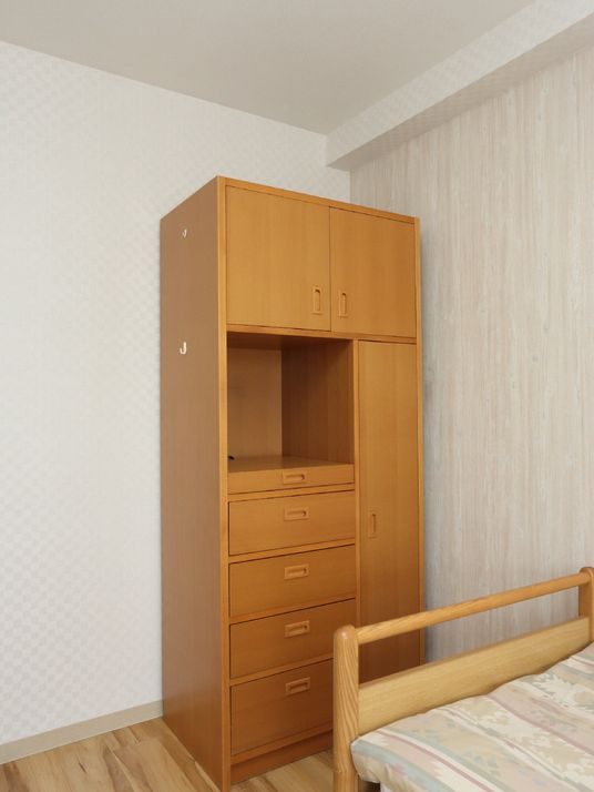 居室のベッドのそばに茶系統のチェストが完備されている。洋服や小物類を収納できるので便利。飾り棚もある。