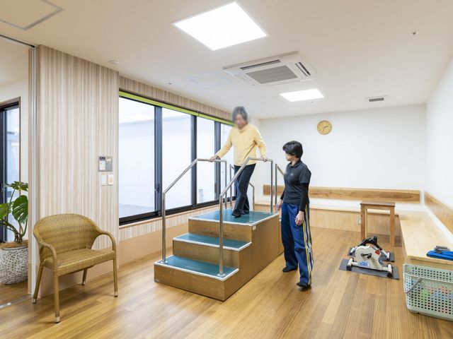 機能訓練の様子 機能訓練室 柔らかな自然光が差し込む空間で、サポートを受けながら階段の訓練が行われています。