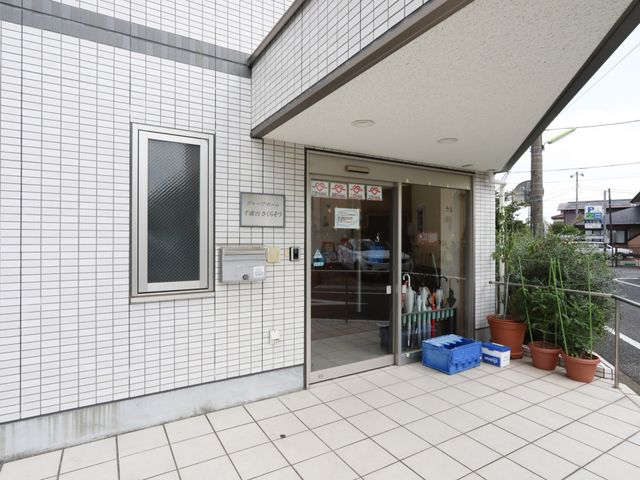白い壁の明るいエントランス 玄関 屋根付きで植物が飾られた入口は、訪れる方々を暖かく迎えます。安全な手すりも設置され、すべての方のアクセスを考慮しています。