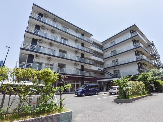 施設の写真 5階建ての建物で、すぐ前に駐車場が完備されている。周囲には背の低い木々が植えられている。各階にはフェンス付きのベランダが設けられている。