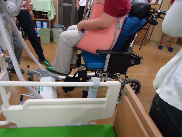 車椅子のトレーニング室 腰に負担をかけない介護教室 スタッフに支えられながら、リハビリ用の器具を使って運動をしている様子です。目の前には、明るく開放的な空間が広がっています。