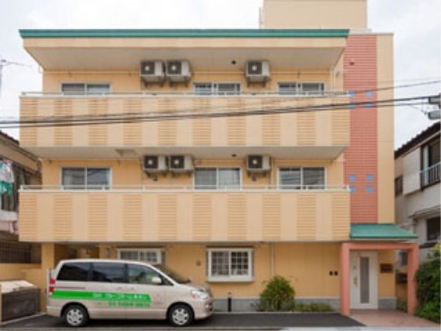 やさしい色合いの建物外観 外観 鮮やかな色合いが印象的な施設です。近くに停車している送迎車が、利用者の方の安全な移動をサポートしている様子が伺えます。