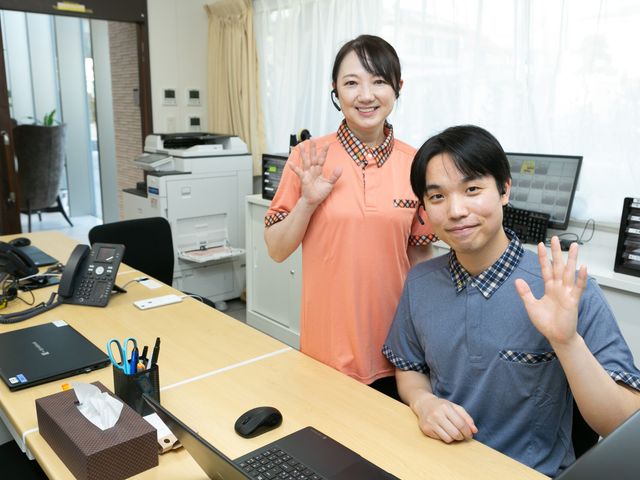 笑顔のスタッフがいる事務室 フロント   明るい事務スペースで、スタッフの方が笑顔で手を振っています。電話やパソコンが整い、来客対応の様子が伝わります。