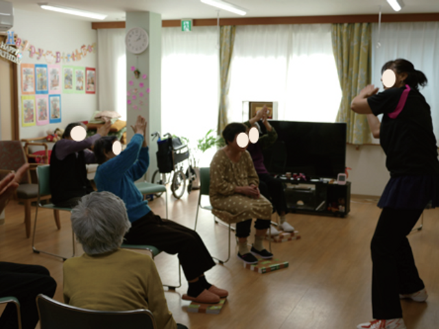 体操をする人々の部屋 機能訓練室 自然光が差し込む明るい空間で、皆が和やかに体を動かしています。机もあるので多彩な活動に利用できます。