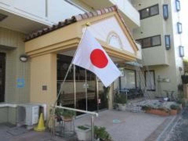 施設の外観入口 外観   正面に日章旗が掲げられた、落ち着いた雰囲気の建物です。出入り口まわりには花があり、親しみやすい印象です。