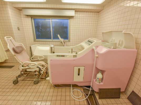白とペールピンクのツートンカラーになっている大きな介護浴槽が設置されている。その左側には、浴室用の特殊な車イスがある。