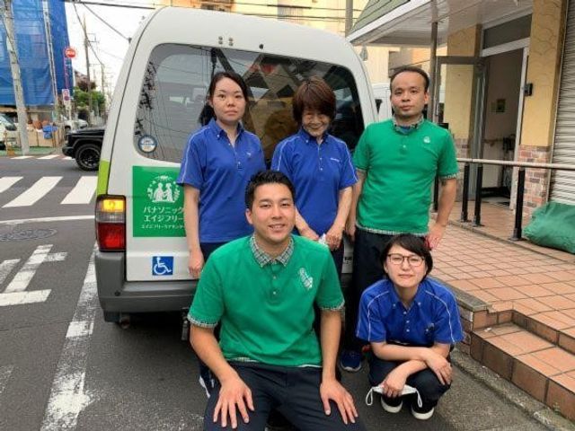 スタッフが車の前に並ぶ外観 外観   送迎車の前でスタッフの方々が並んでいます。青と緑の制服が見え、就業時の雰囲気をイメージしやすいです。