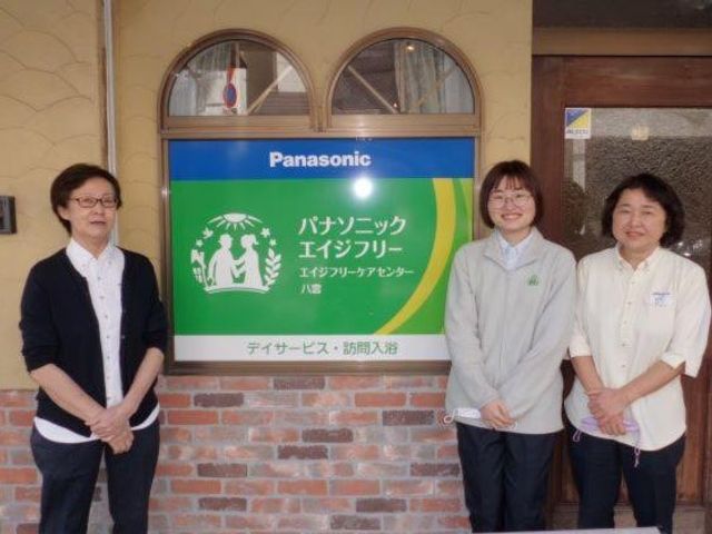 スタッフが並ぶ看板前の外観 外観   施設の看板の前に3人のスタッフが並んで立っています。入口まわりの落ち着いた雰囲気が伝わり、働く場の印象を確認しやすいです。