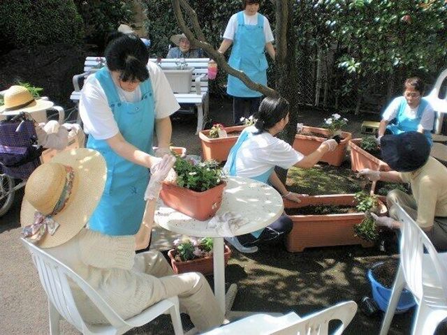 屋外で園芸活動をする人々 レクリエーション 青空の下、利用者は笑顔で園芸作業に興じています。スタッフの支援を受けながら、心地よい時間を過ごせる環境です。