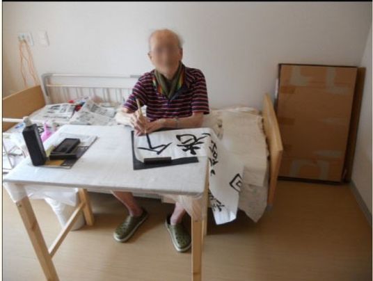 居室で撮影された写真。男性入居者様が介護ベッドの上に座って、習字を楽しんでいる。新聞の上に墨汁が置かれている。