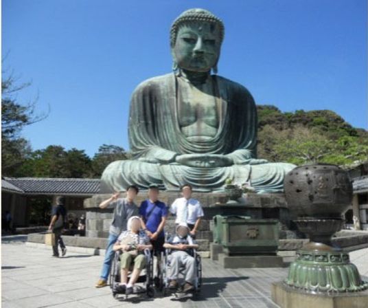 天気の良い日に大仏前で撮影された写真である。車椅子に座った２人の入居者様の後ろに男性が３人立っている。